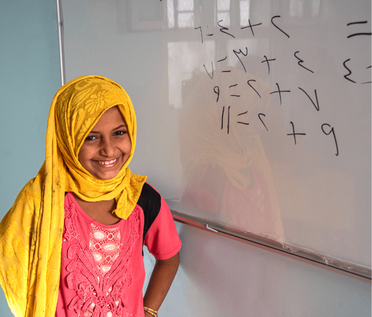 girl in a classroom standing in front of whiteboard smiling