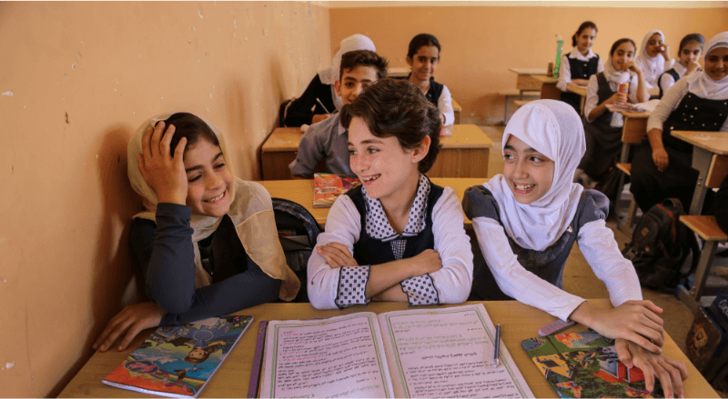 Three students in their classroom sitting at their table smiling and laughing together