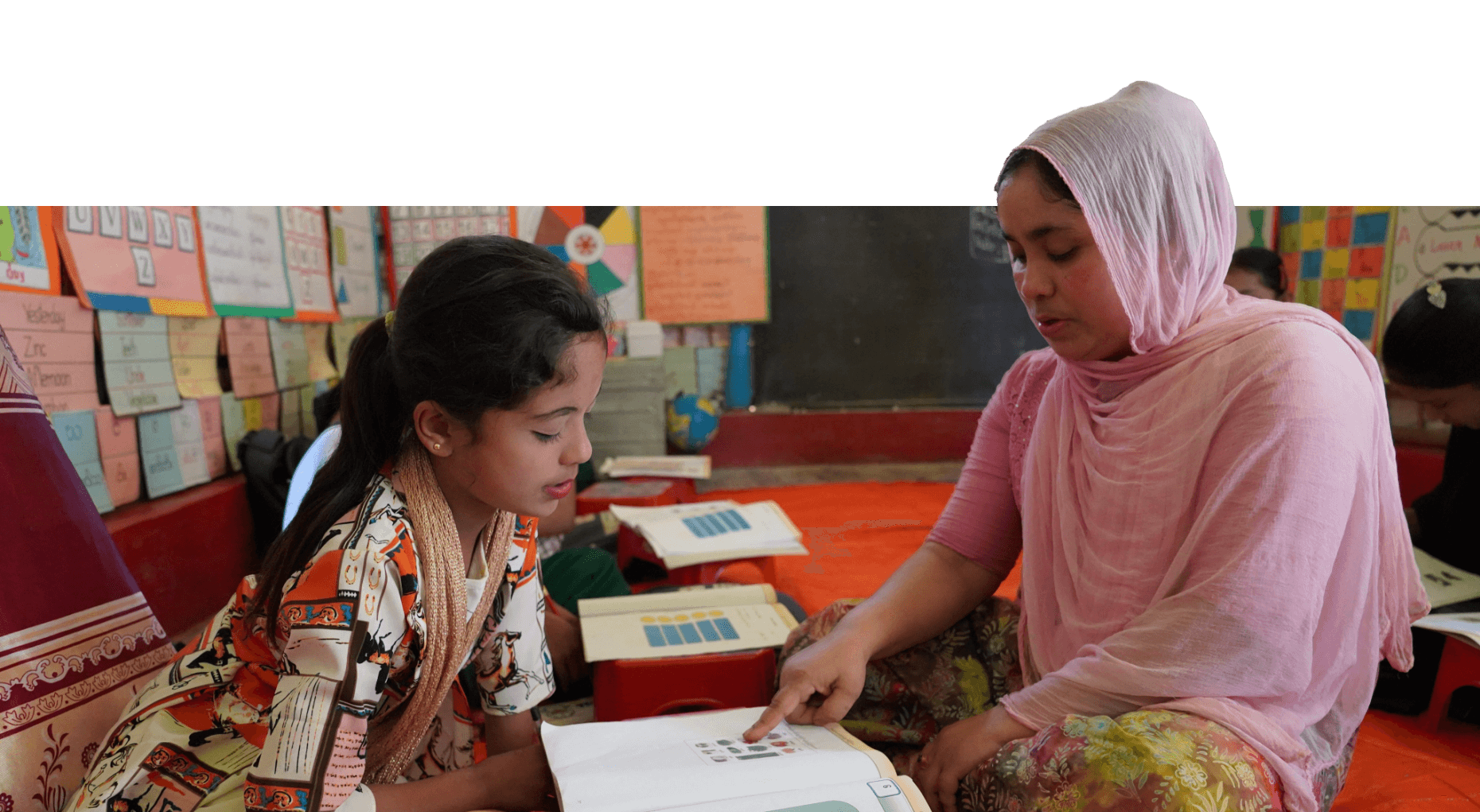 a student sits with her teacher on the floor at school. They are looking at a workbook together.