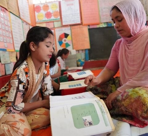 a student sits with her teacher on the floor at school. They are looking at a workbook together.