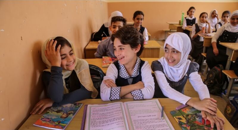 a student sits with her teacher on the floor at school. They are looking at a workbook together.