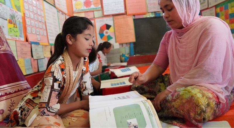 a student sits with her teacher on the floor at school. They are looking at a workbook together.