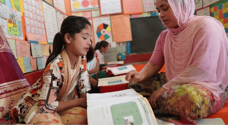 a student sits with her teacher on the floor at school. They are looking at a workbook together.