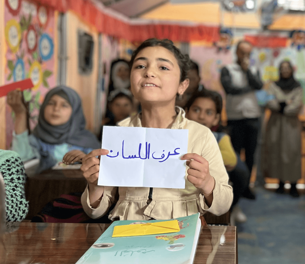 A studnent in her classroomo holding up a piece of paper with Arabic words written on it
