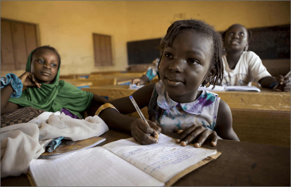 A girl in her classroom with her classmates. She is writing in a notebook with a pen.